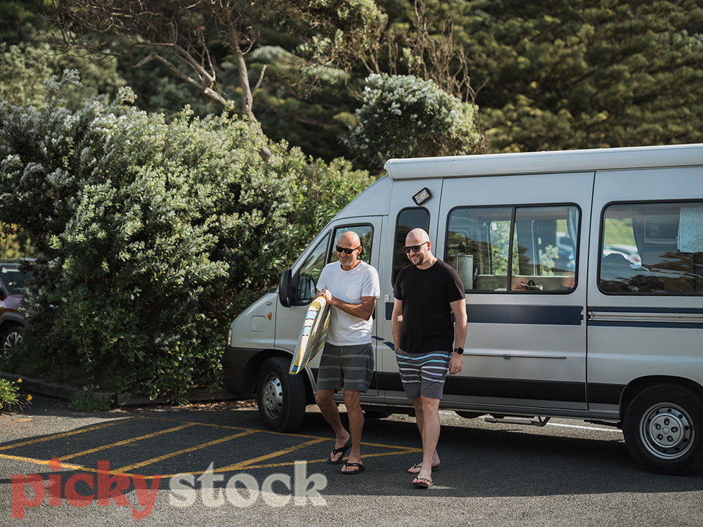 Two mean, one holding surf board, walking in beach clothes towards camera. Caravan behind them