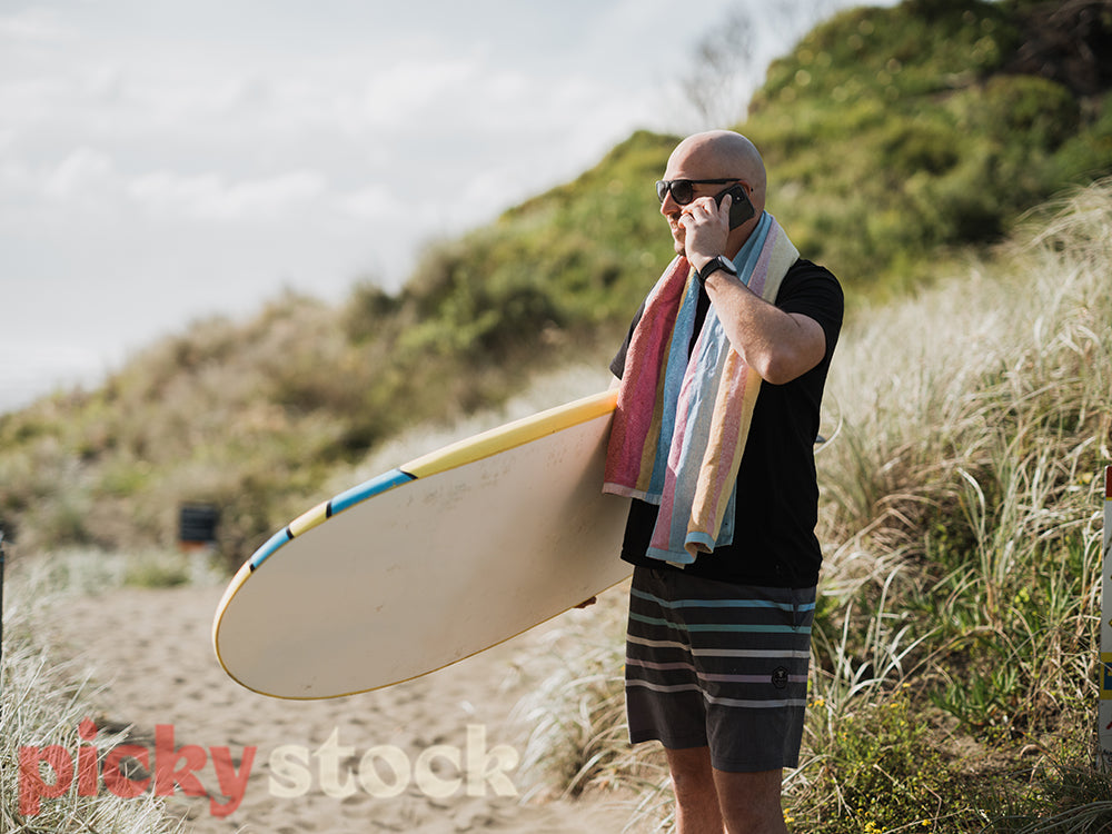 Man walking down to beach wearing black top and holding surf board talking on mobile phone.