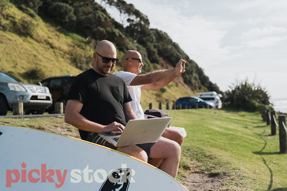 Man sits by beach working on laptop. Other man sitting next to him taking photo