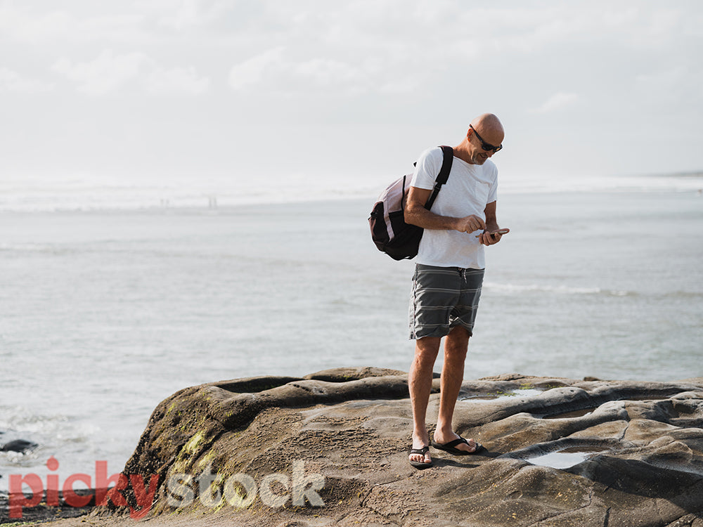 Man standing on rock by ocean looking down at mobile phone