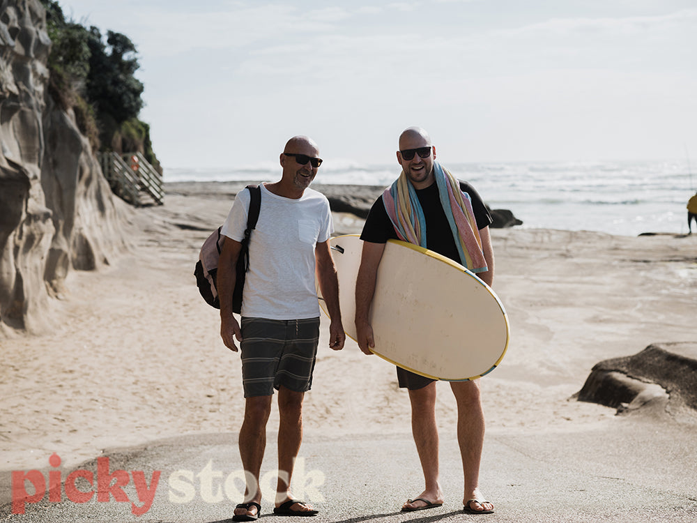 Father and son standing on coastal sand path looking at camera. One man holding surf board