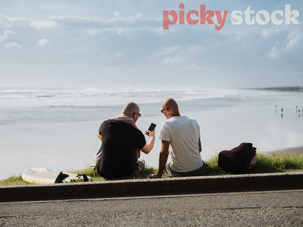 Two men sitting on grass towards ocean. both men looking down at one mobile device.