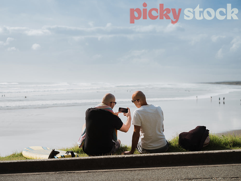 Two men sitting on grass towards ocean. both men looking down at one mobile device. One man pointing at screen