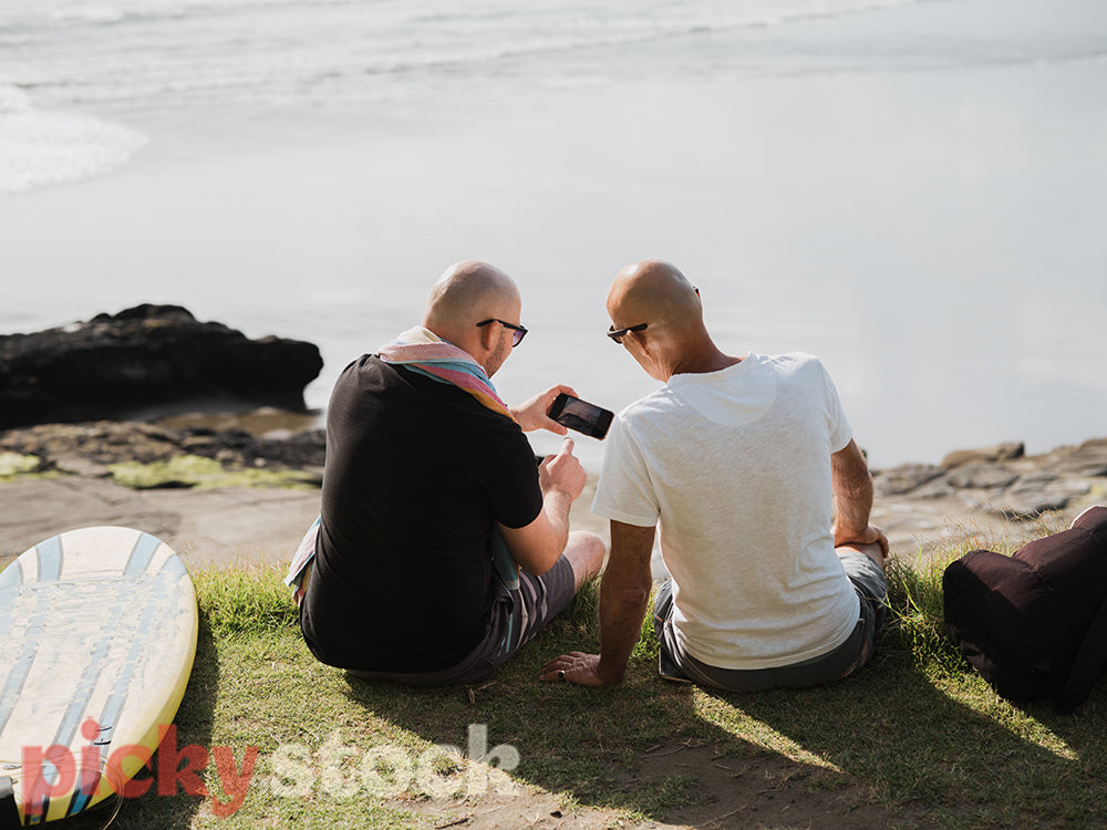 Two men sitting on grass towards ocean. both men looking down at one mobile device. One man pointing at screen