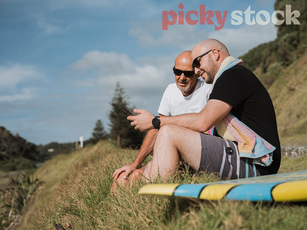 Two men sitting on grass towards ocean. both men looking down at one mobile device.