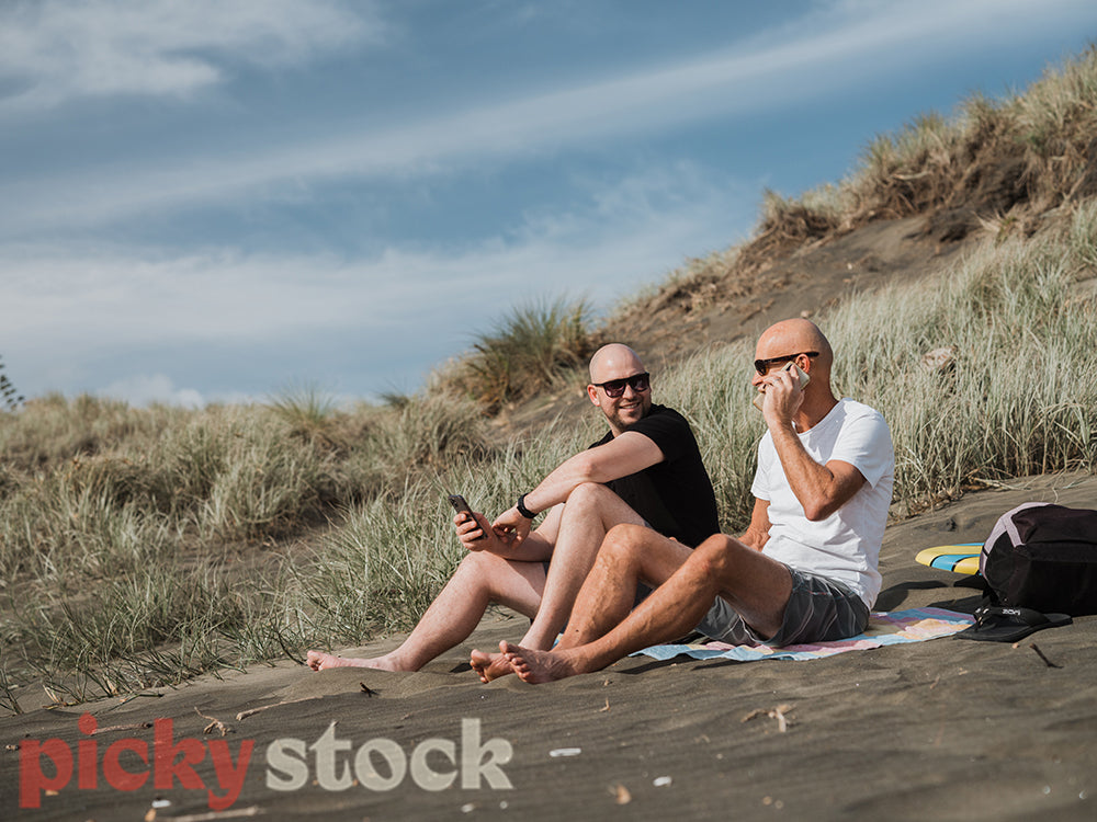 Two men sitting on sand with dunes behind them. One man on phone call