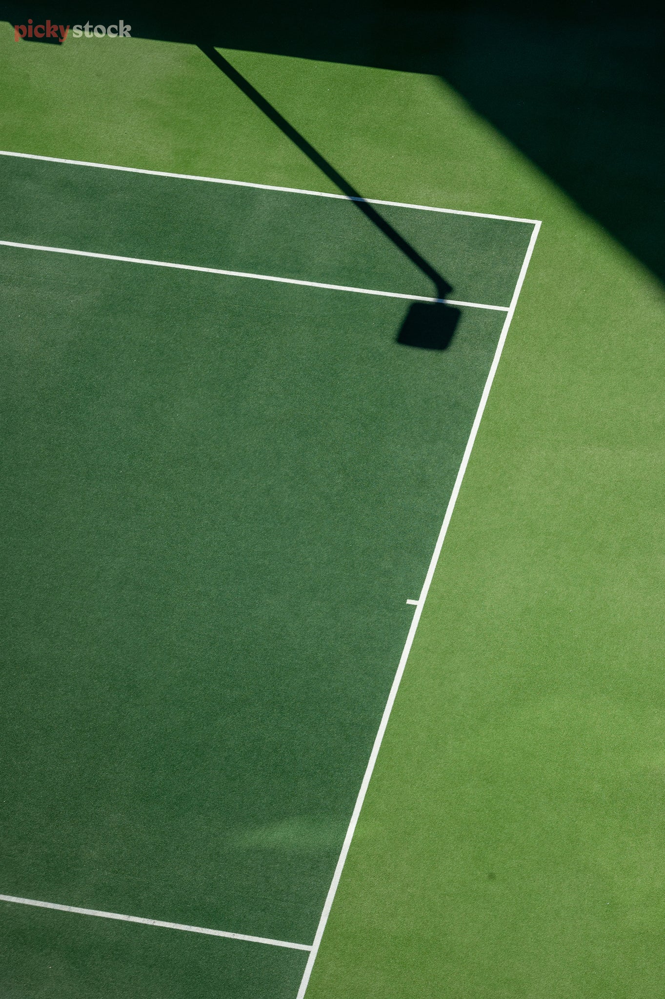 Looking down at the corner of a astroturfed tennis court. The different colour greens split by the white lines of the court, with graphic angular shadows from the surrounding wall and court lights hitting the ground. 