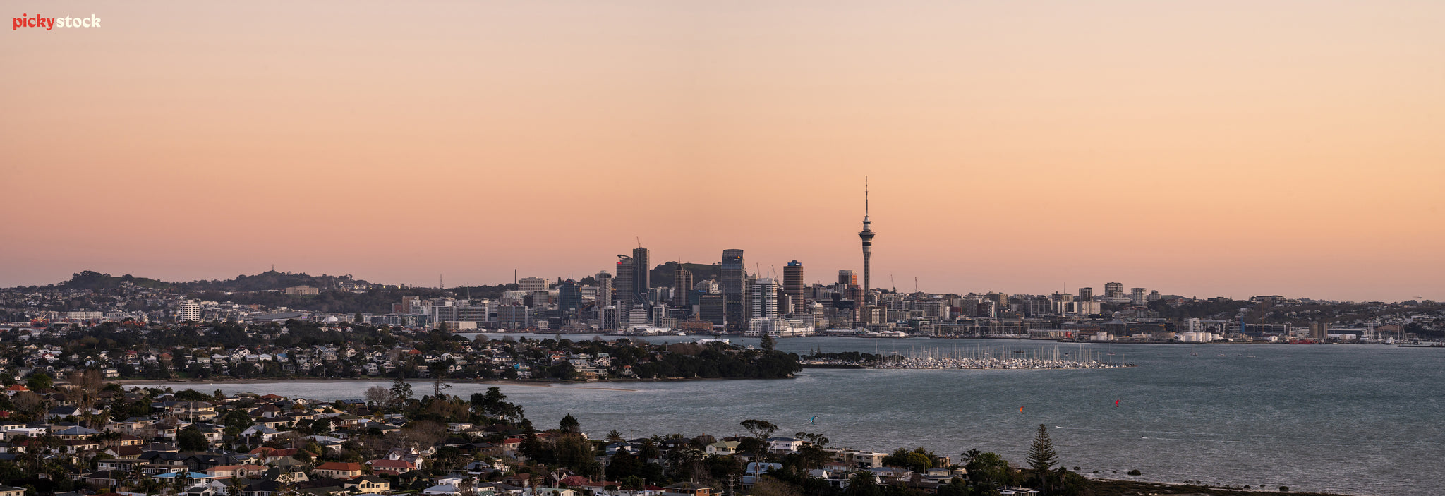 Panorama of Auckland City at sunset, as seen from a higher viewpoint than the city. The sky is changing to a pinky / orange as there are no clouds in sight. 