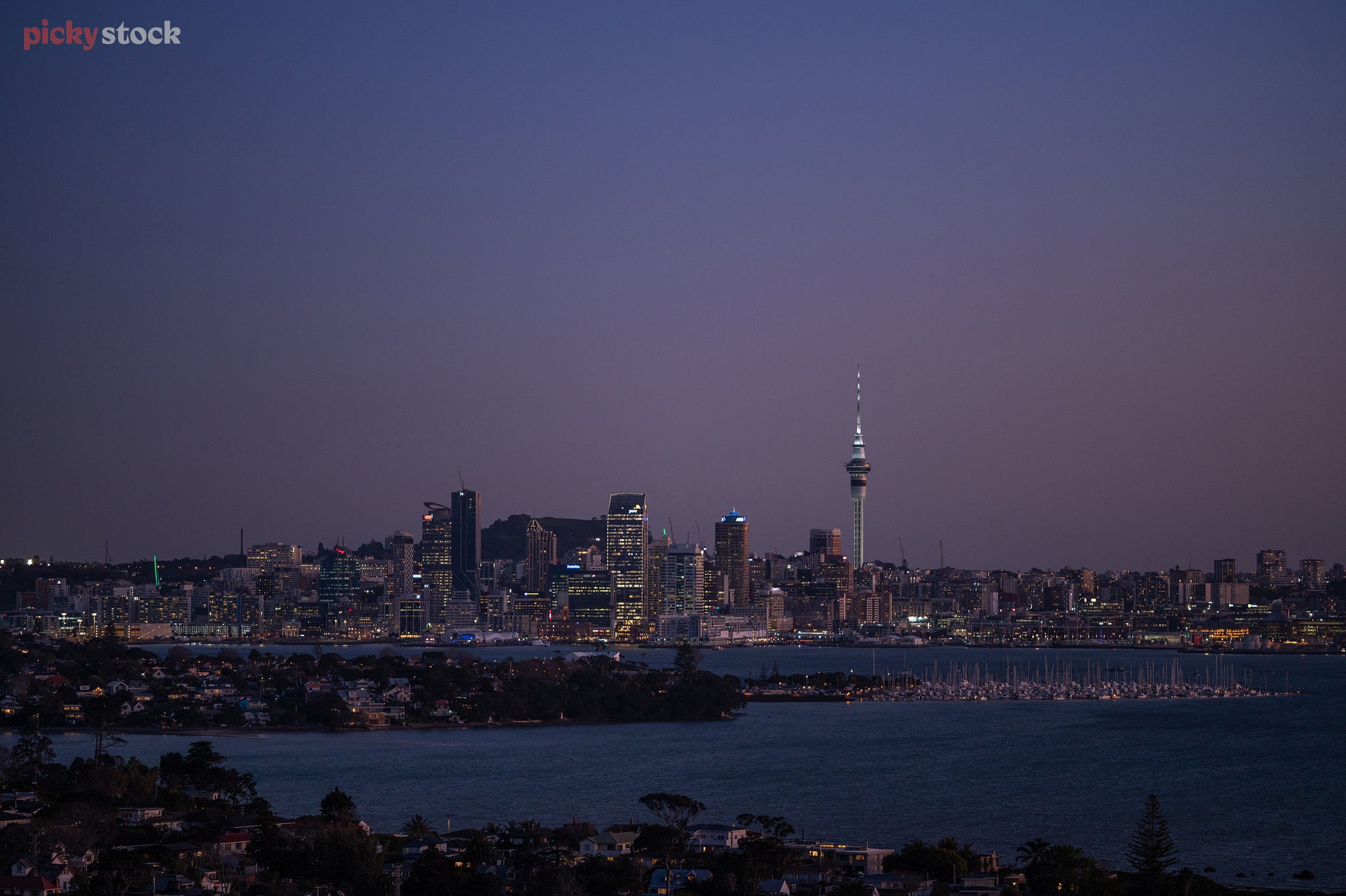 Wide shot of Auckland City just as night hits, there are few lights lighting up the skyline as the dark buildings hit the deep blue clear sky. 