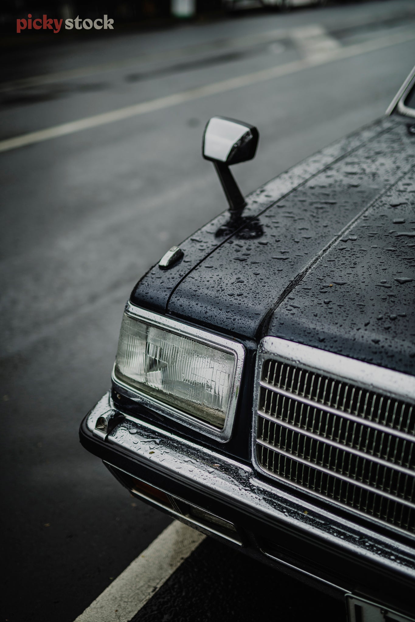 Droplets of water sit on the bonnet of a vintage navy / teal car. Just the right side of the car is visible with the front grill, wing mirror and headlight as it sits on an angle against the wet ashfelt.