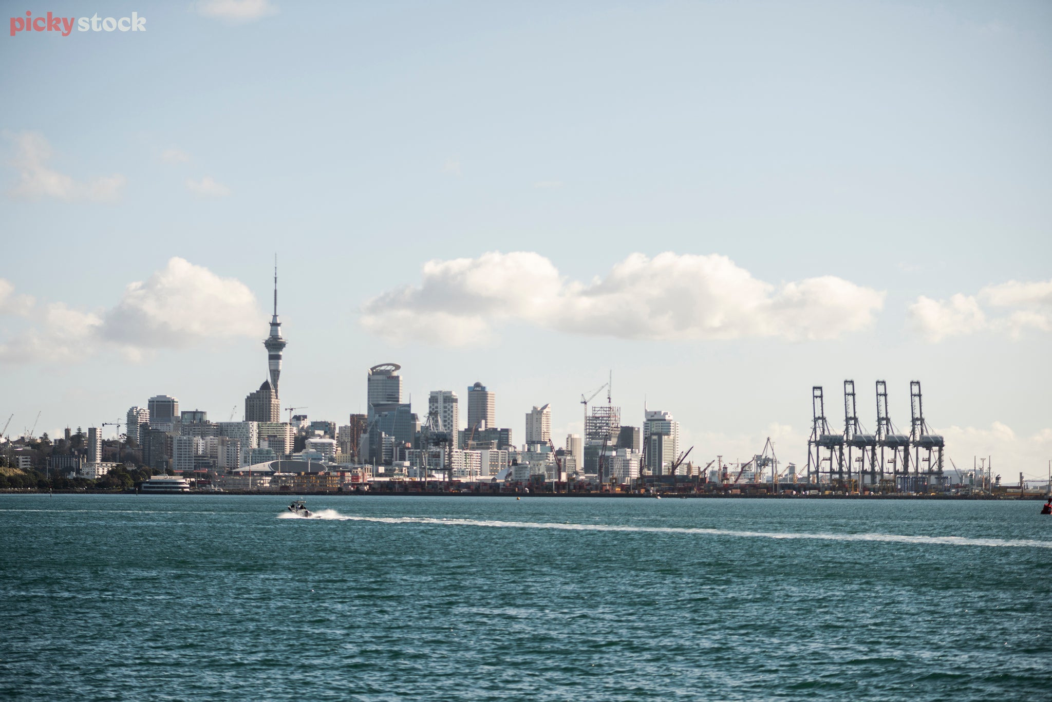 A light blue sky fades to white at the horizon. Small clouds float across. The cities skyline reaches up into the sky. On the left the sky tower, tall and thin extends highest. On the right a port filled with containers and cranes it's darker than the city. The city and port sit on the harbour, a blue body of water which fills down to the foreground. from right to left a small boat is motoring, leaving a white wake churned in its path.