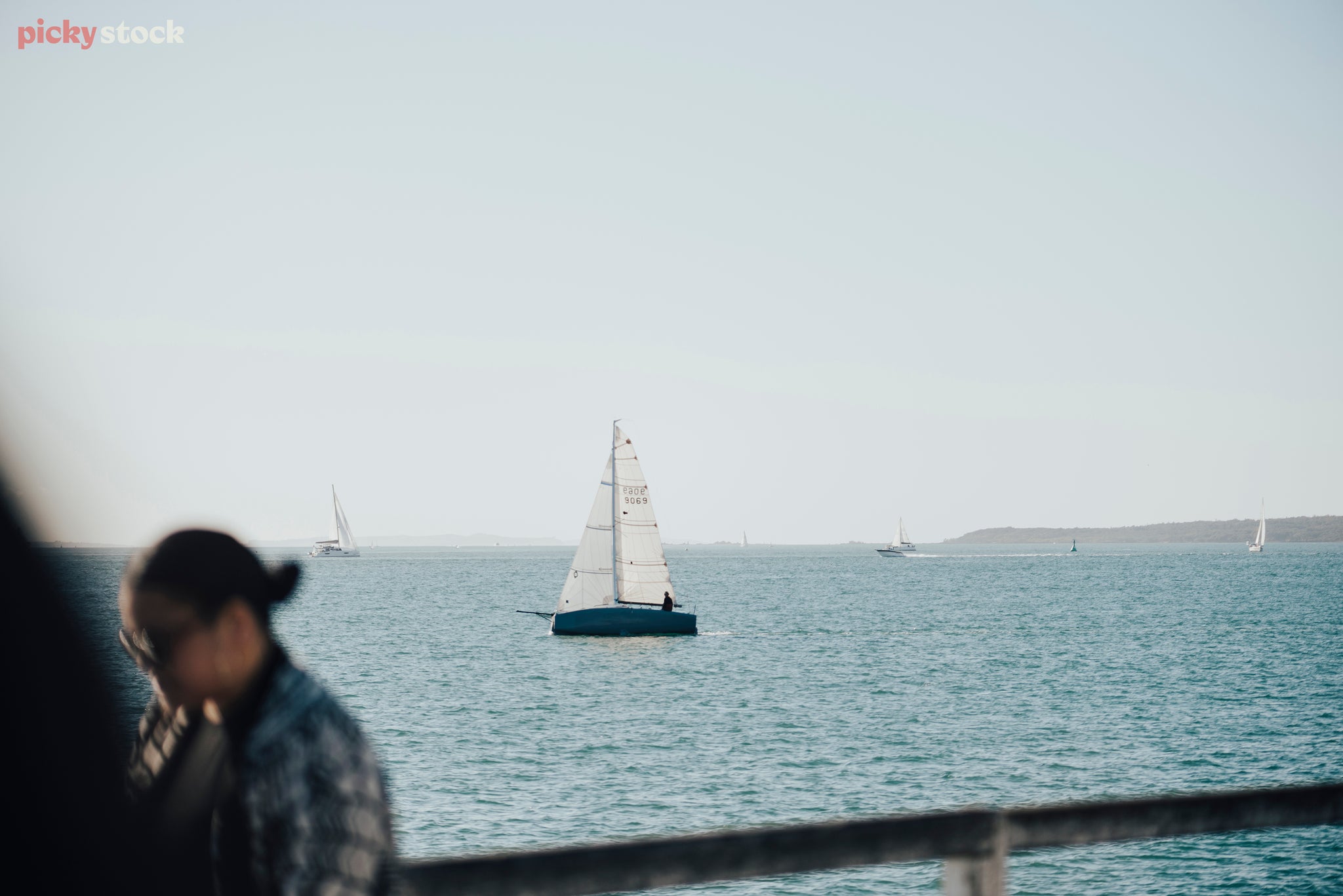 A cloudless light blue sky turns almost white at the horizon. In the distance on the left a finger of land sits on the water, around it small sail boats ply the waters of the Hauraki Gulf.
Closer to frame a lone sailboat catches the wind and as a single sailor guides it through the water. In the foreground is an out of focus woman and hand rail.