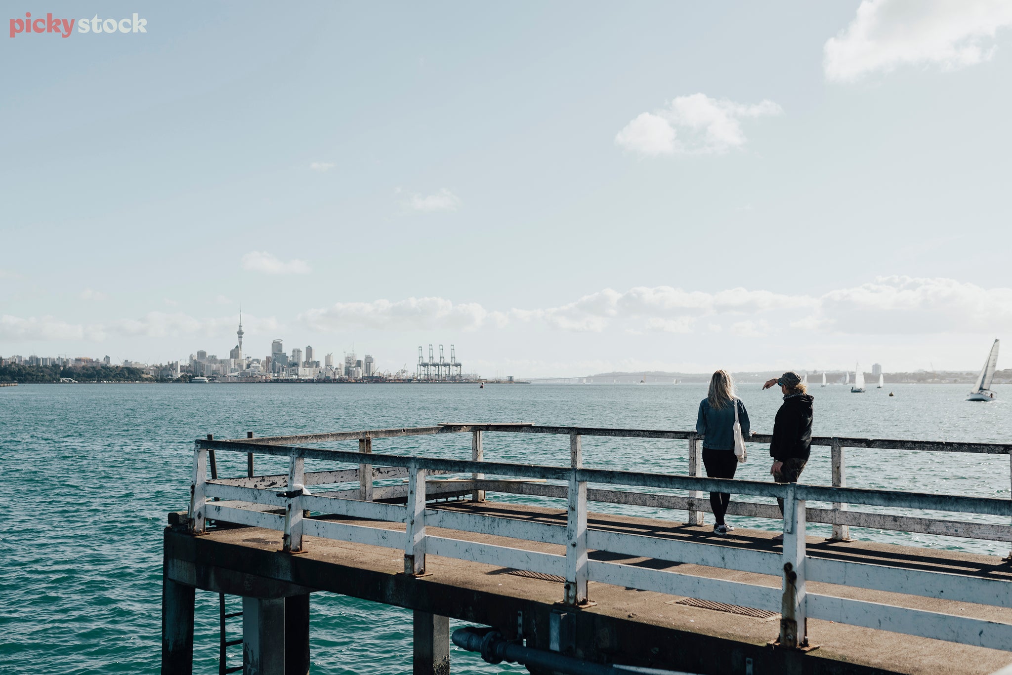 Two figures lean over the railing on the fishing wharf, looking back to Auckland on a blue day. 