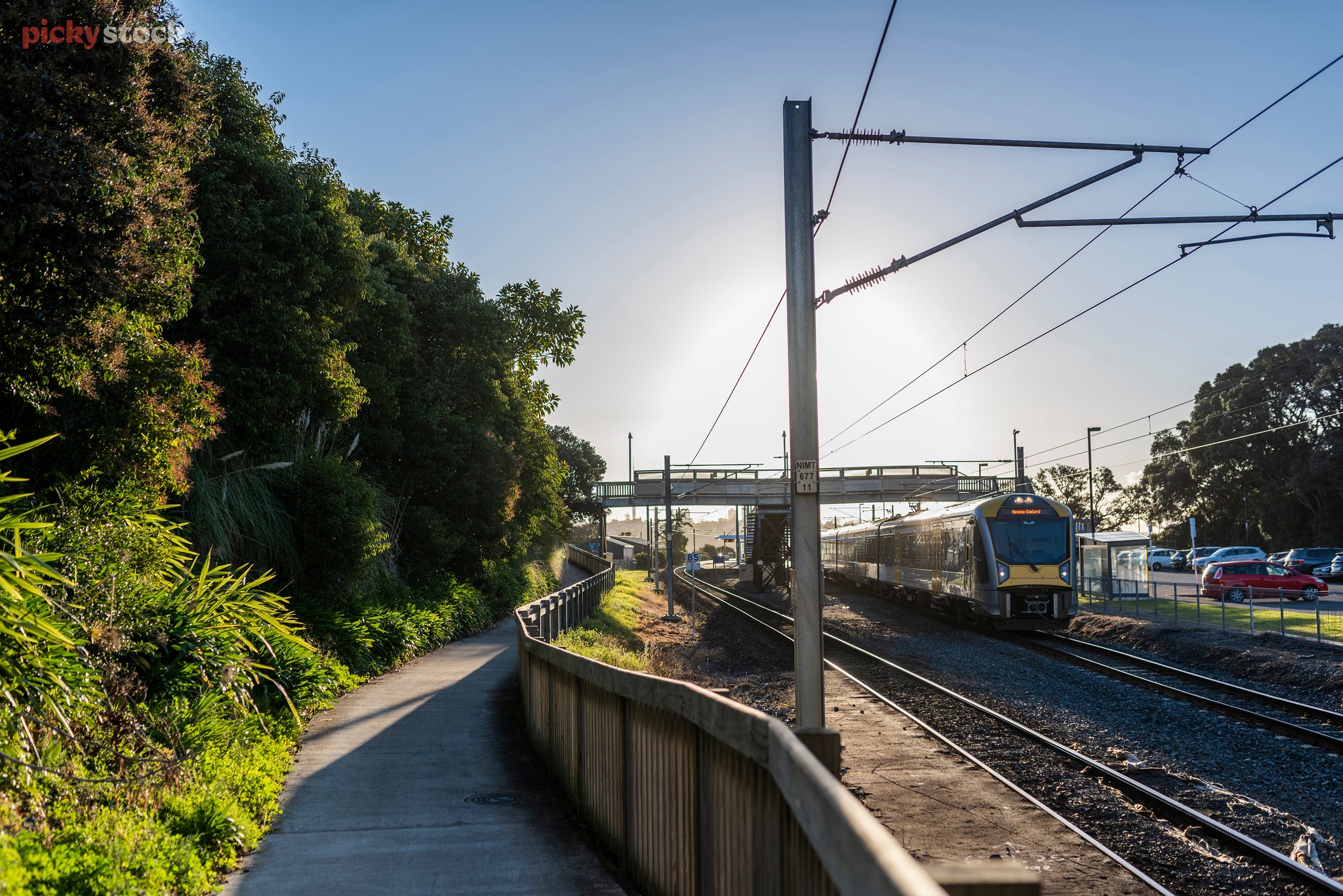 Early morning blue sky becomes white on the horizon where the sun is rising. On the right a train has stopped at a station. There is electric cables strung above the train and the tracks. On the left a bank has trees and flax following a concrete walking path and wooden fence. The path leads up to an over bridge across the tracks and cables. Giving access to the train platform. On the far right cars a parked. 