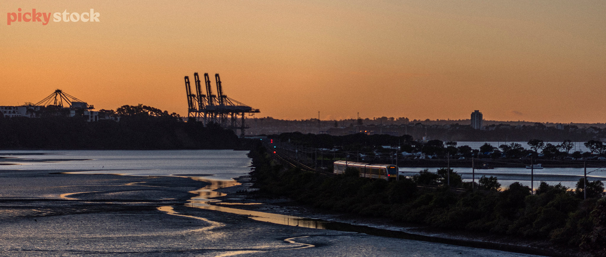 A train track cuts its way across a tidal esturary towards a port where 4 large container cranes sit idle.