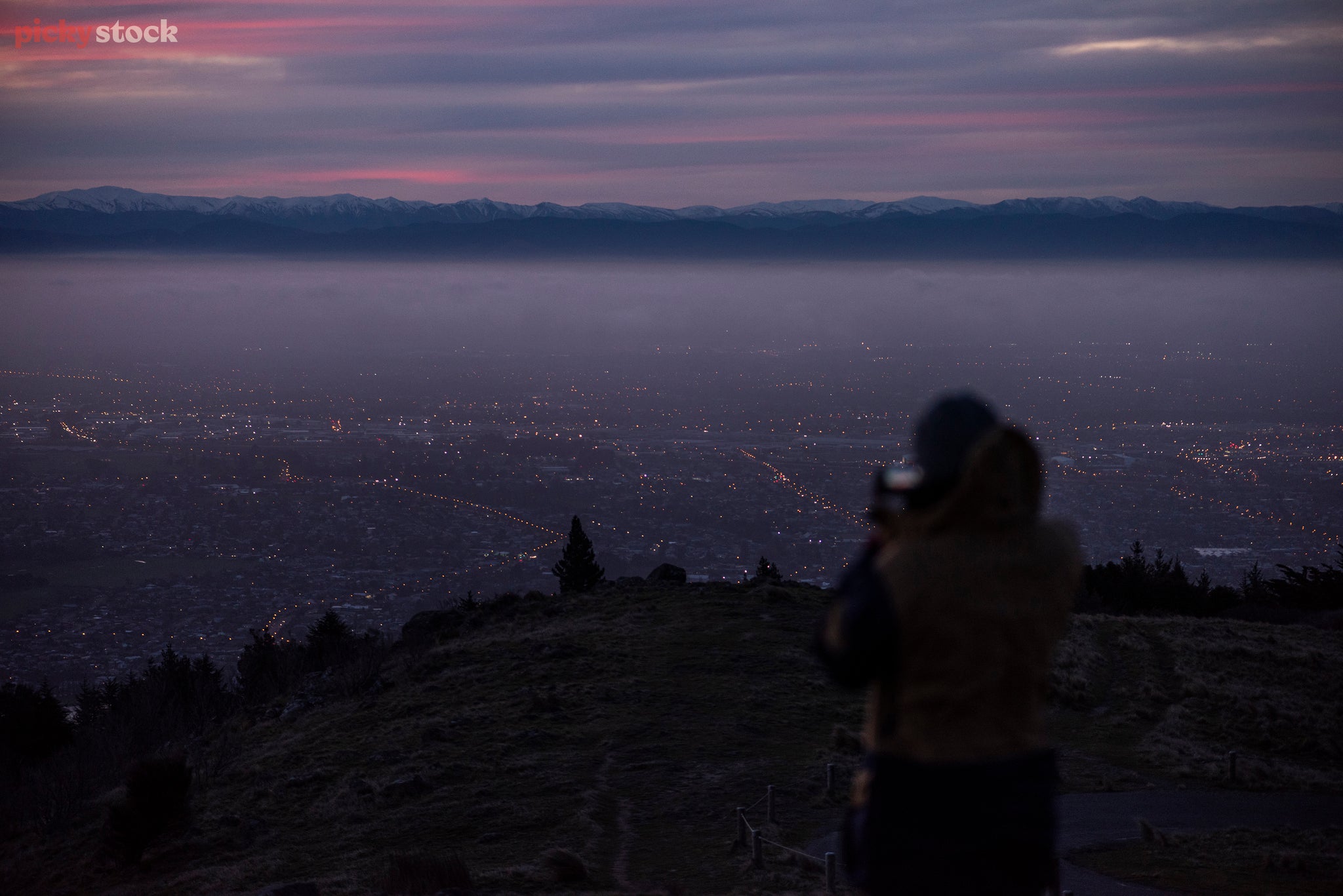 A very hazy and dark viewpoint from the Moody Port Hills overlooking Christchurch. In the far distance the purple and pink dark sunsetting just breaks up the night sky. The silhouette of a person breaks the scene to the right. 