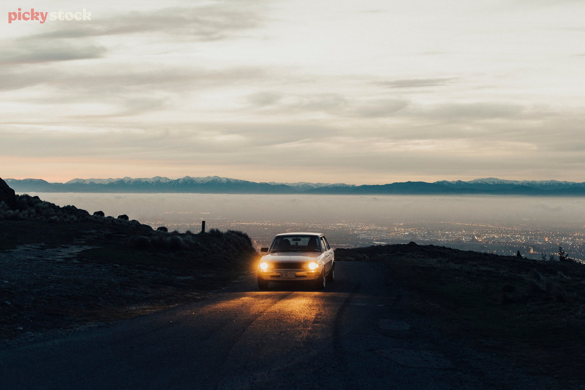 Car with lights on drives towards the camera, in the distance snowy mountains cut the horizon through the white cloudy sky.