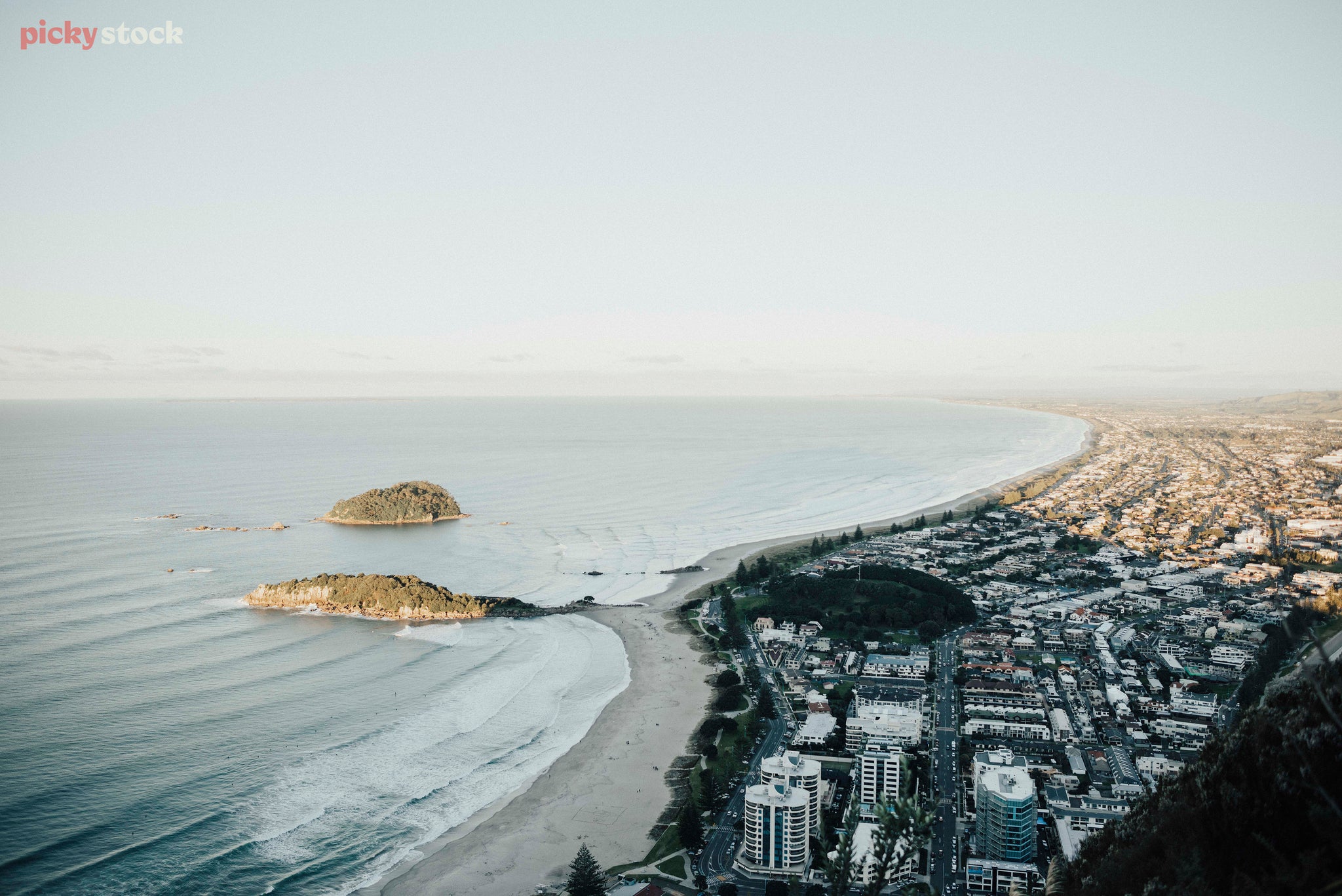 Ariel view of the Mount in Tauranga, a township built on a beach. There are two islands and waves which break on shore.