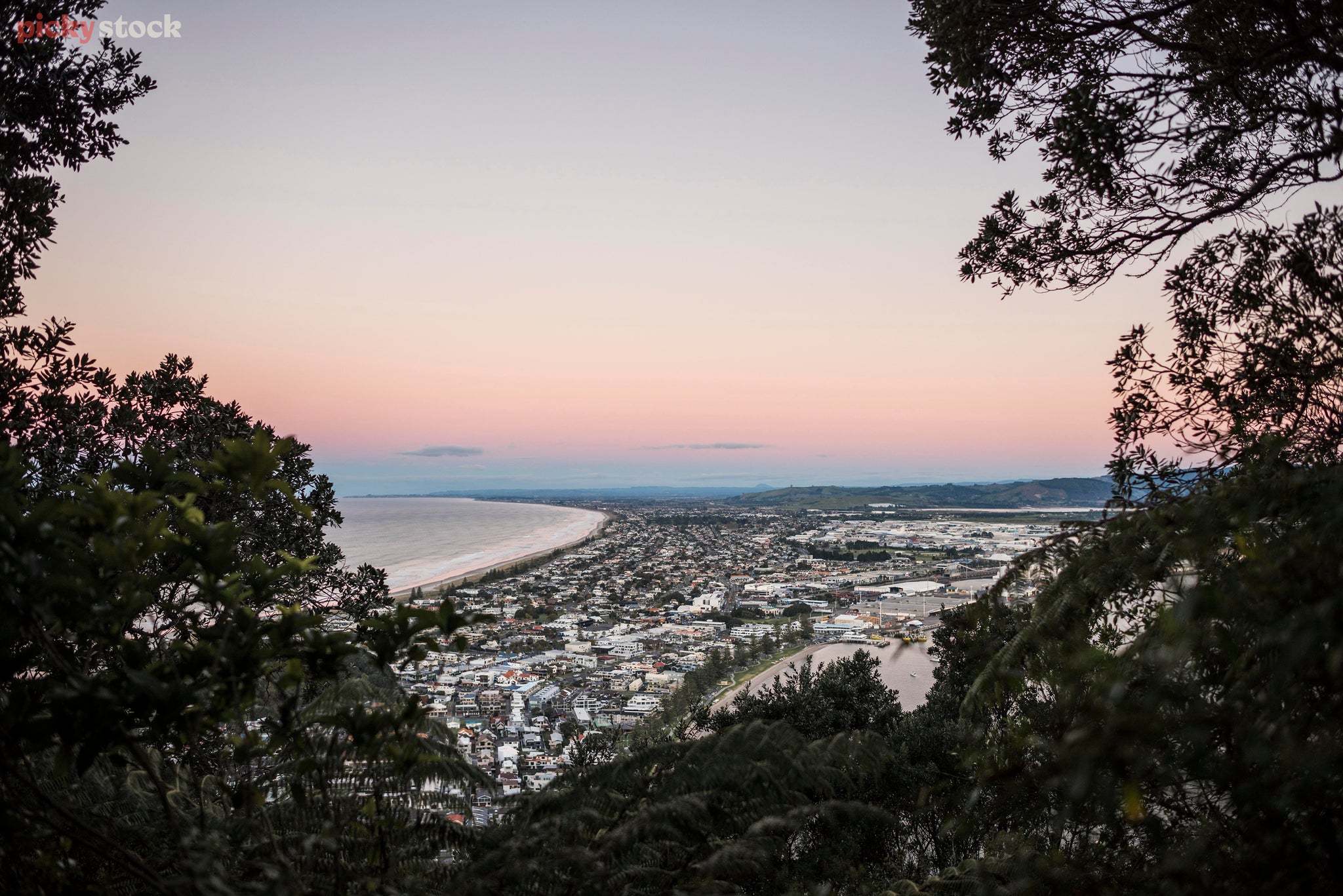 A view of the Mount in Tauranga New Zealand at night, the pink sky hangs above the town built on a beach and next to a port.