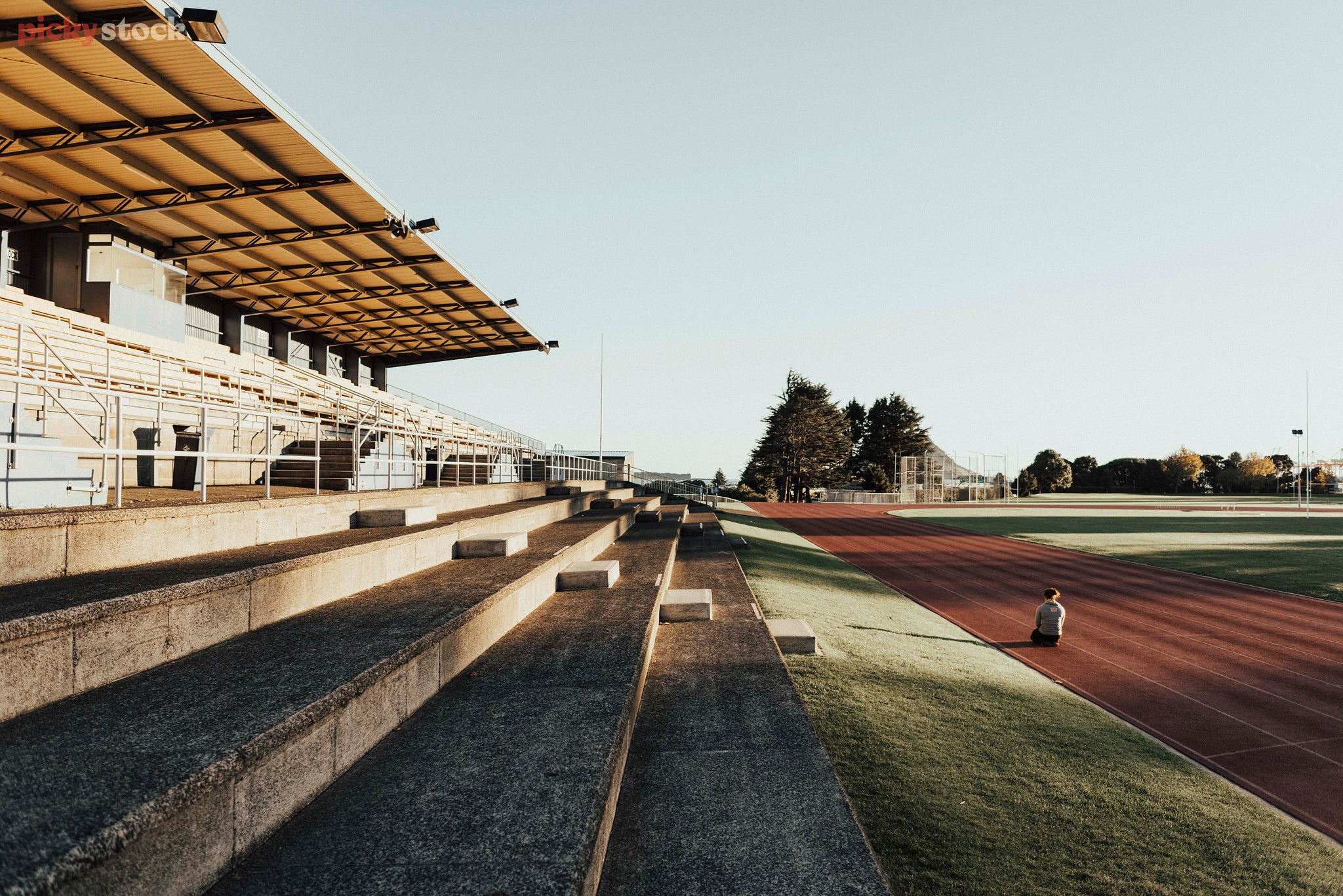 Young woman sits with her back to us on a running track in the early morning. Next to the track is a large stadium seating building.