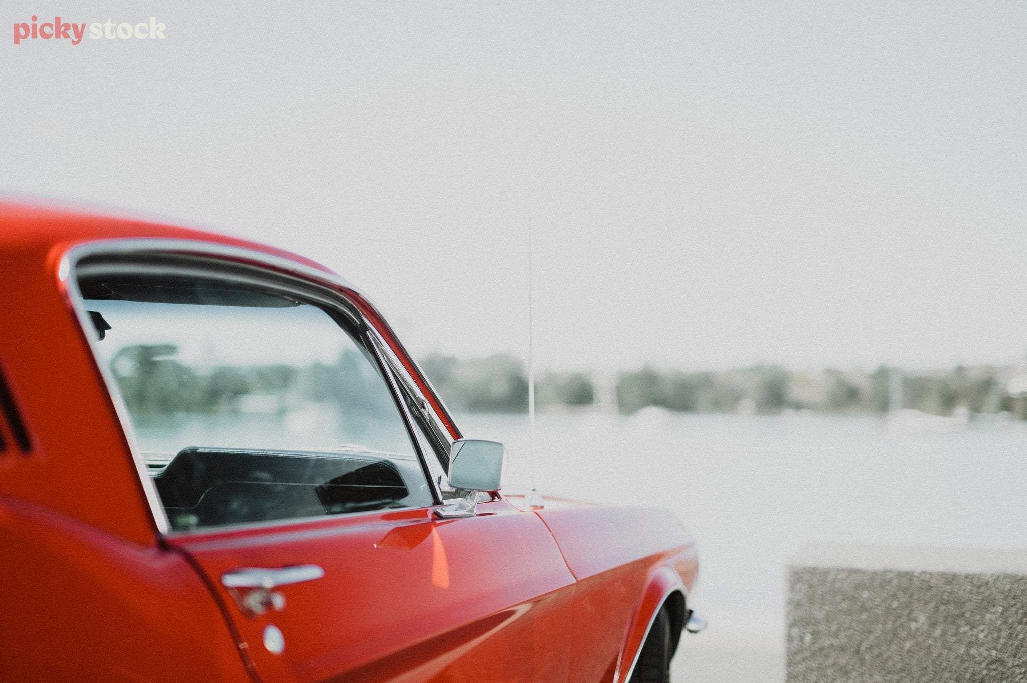 The right hand side of a red Mustang sports car over looking an out of focus harbour.