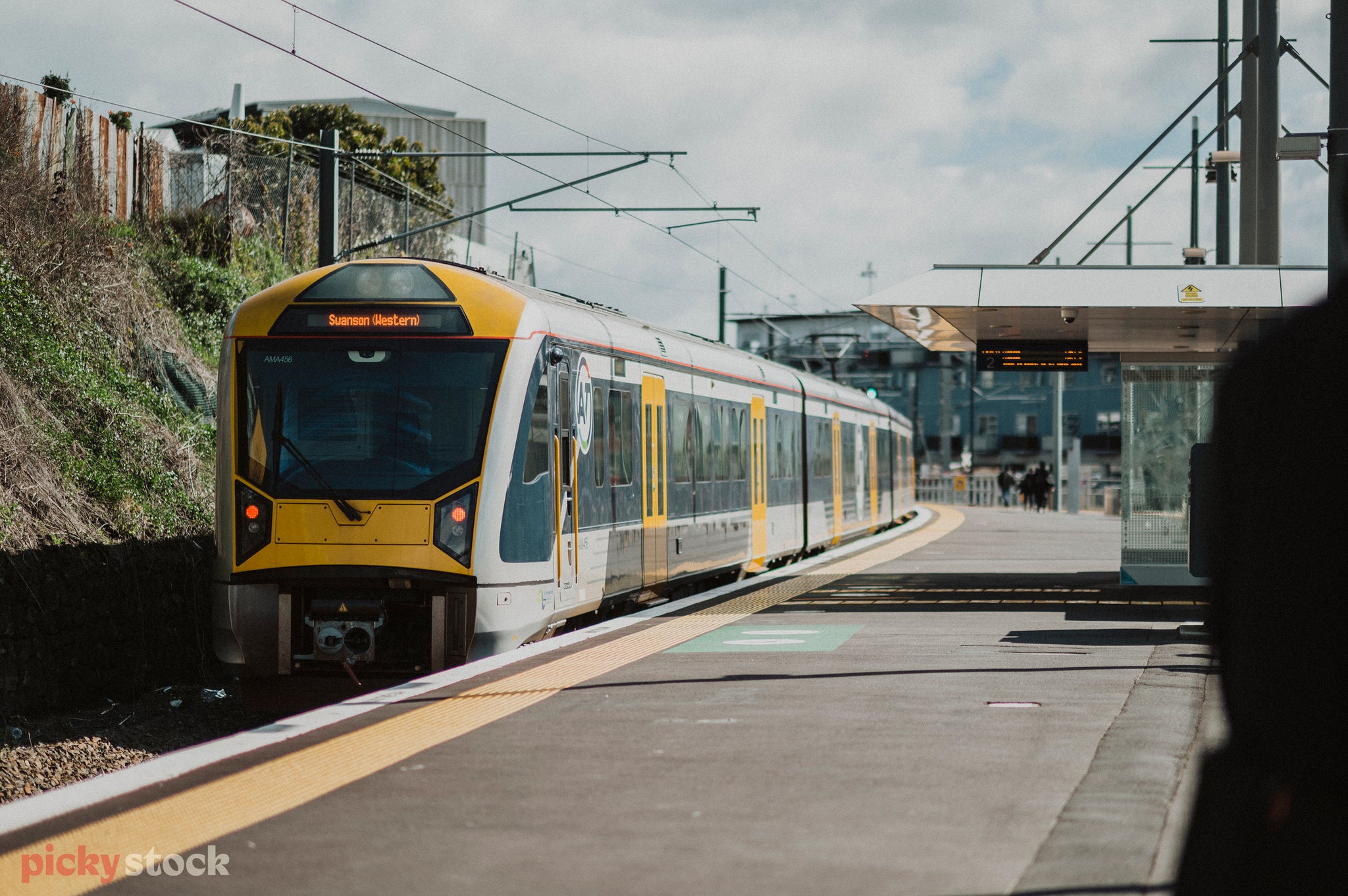 A grey and yellow electric train slides into the station at Mount Eden, hard to tell what time of the day. A shadowed but friendly figure stands to the right of frame. 