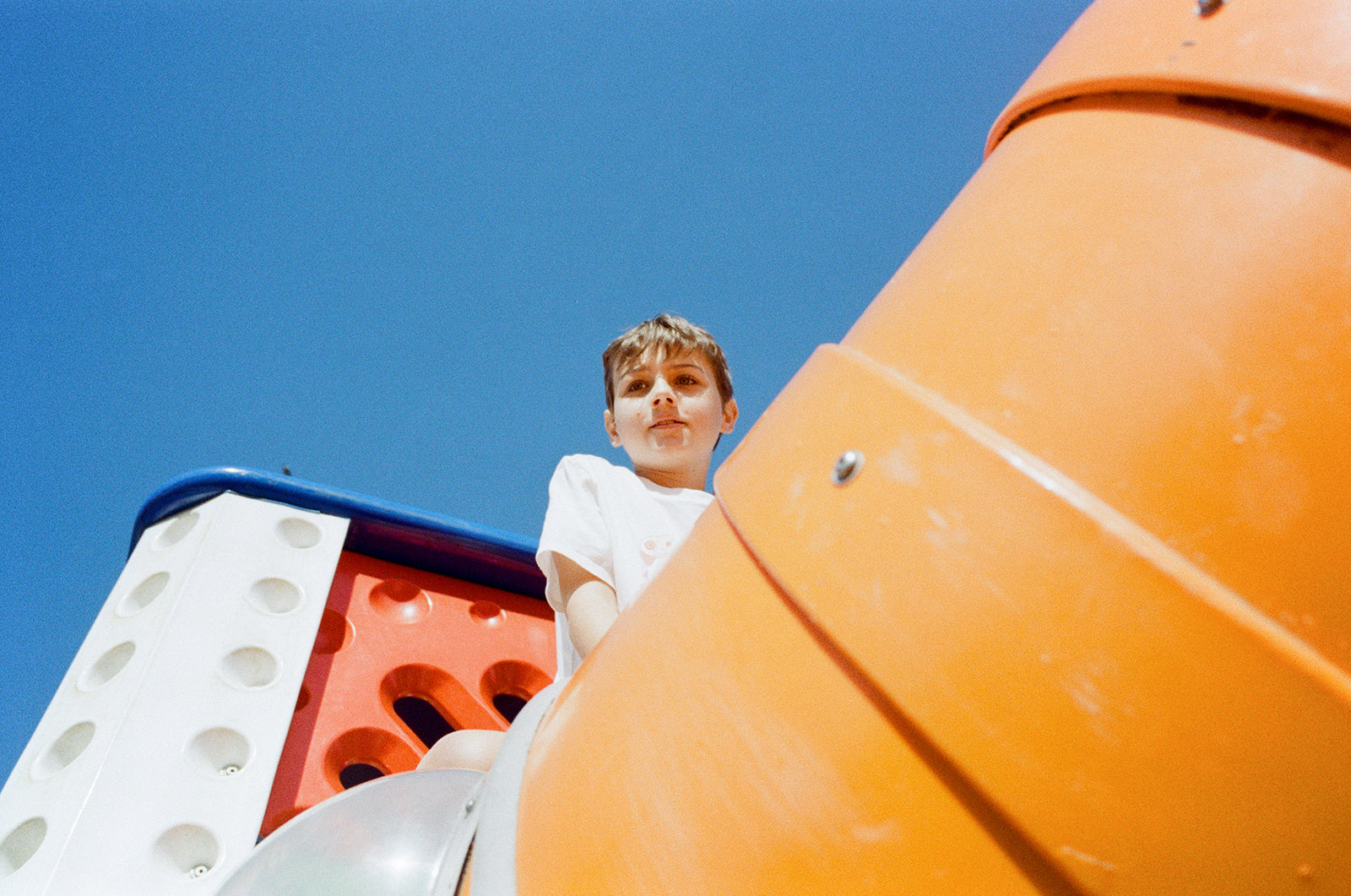 Little boy sits atop an orange tunnel slide at a sunny playground.
