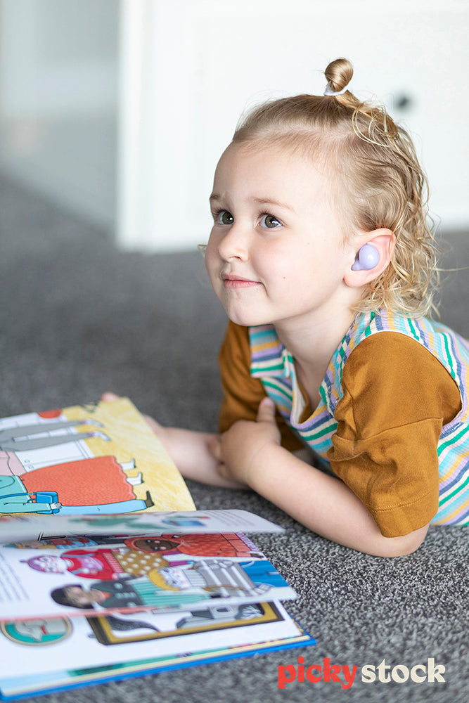 Young blonde girl lying on the ground, reading brightly coloured picture book. Wearing purple air pods looking up off camera 