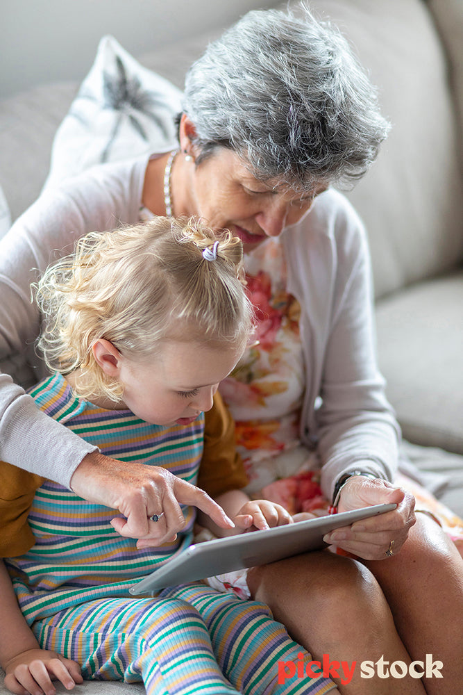 Portrait image of young girl sitting on couch with grandma. Nana is pointing at screen showing small girl something on her silver ipad tablet device. Girl is watching on wearing stripped colour outfit 