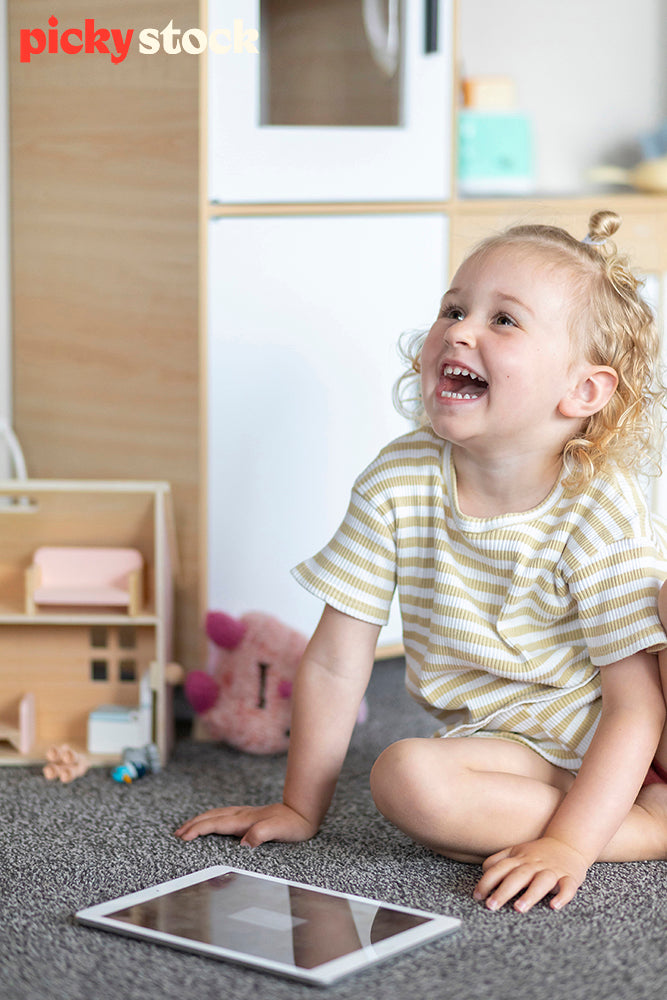Portrait image of girl playing with ipad on the carpet in her bedroom. She is looking up with a huge smile on her face. Little dolls house in the background. 