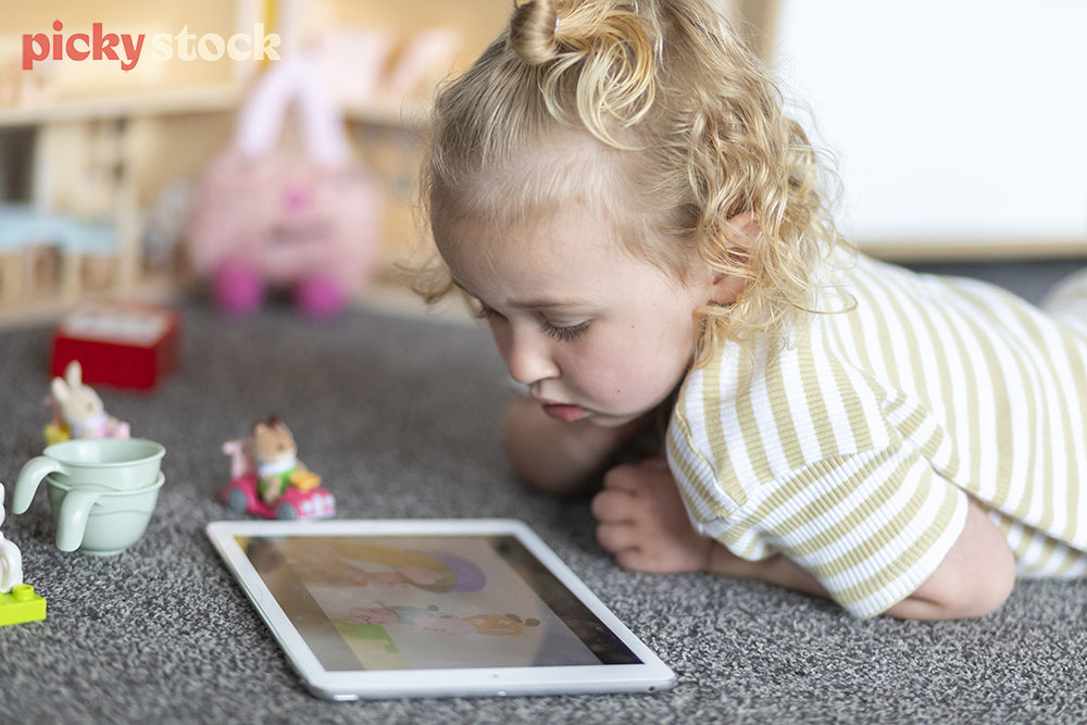 Landscape image of a young girl playing with ipad on the carpet in her bedroom. Little dolls house and kids toys in the background. 
