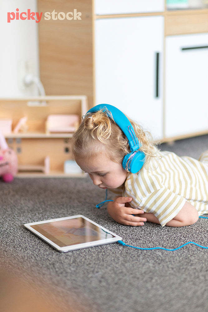 Portrait image of a young girl weaering bright blue headphones playing with ipad on the carpet in her bedroom. Little dolls house and kids toys in the background. 
