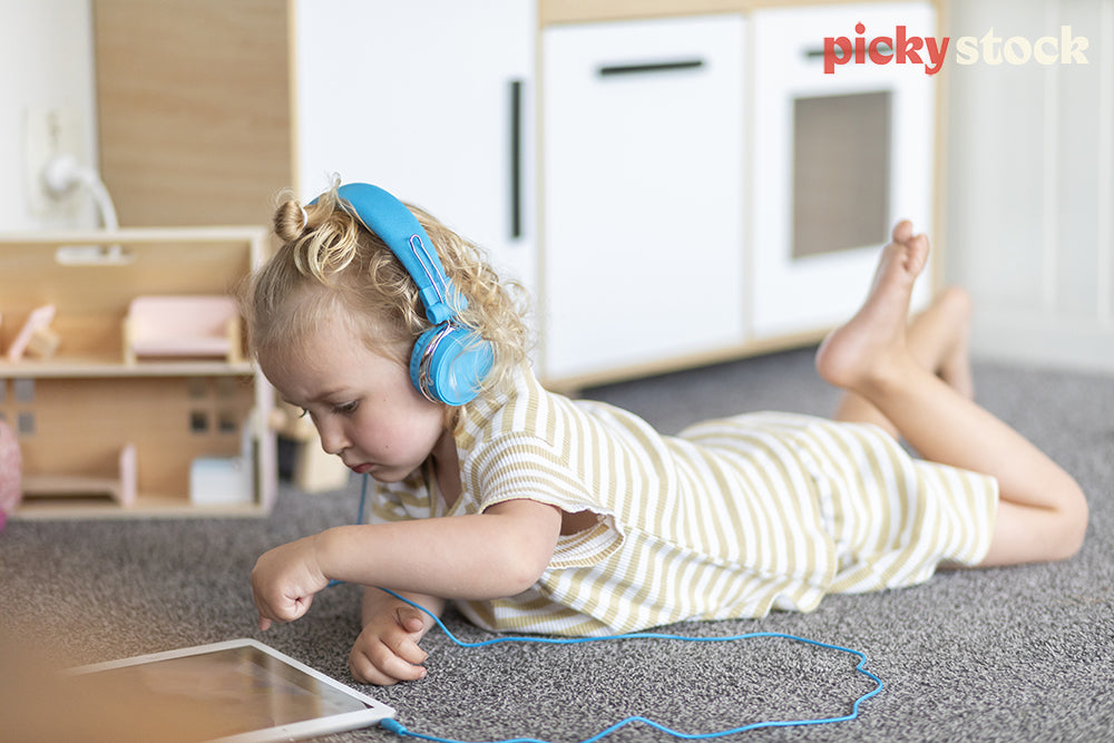 Landscape image of a young girl wearing bright blue headphones playing with ipad on the carpet in her bedroom. Little dolls house and kids toys in the background. 