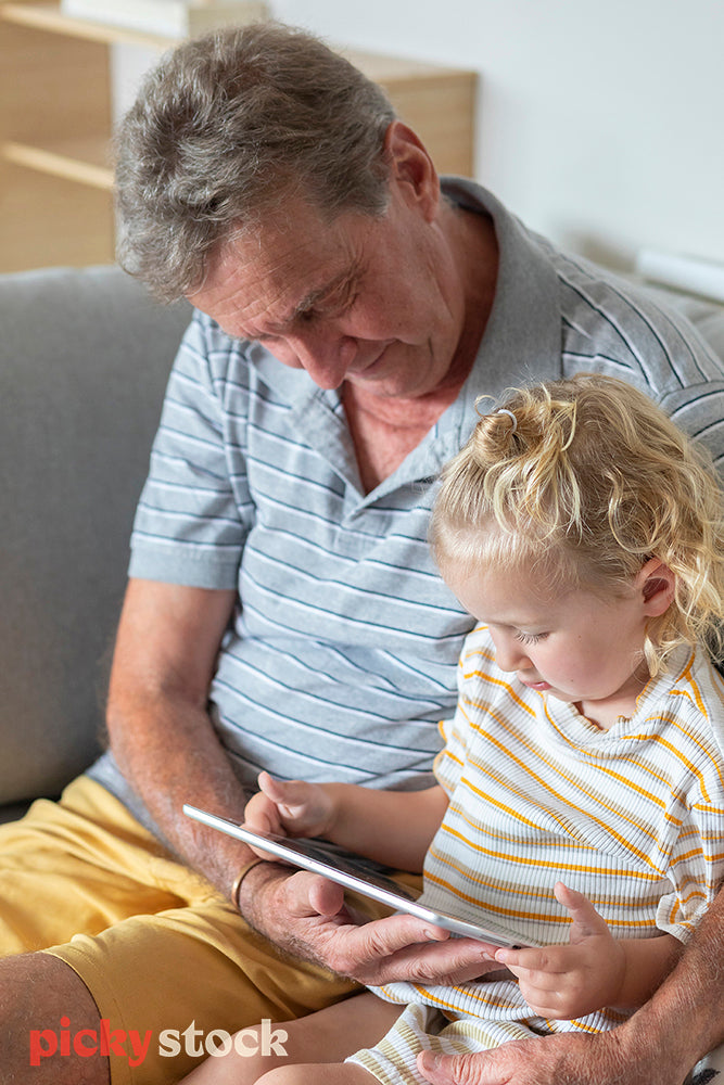 Young girl sitting next to grandfather on the couch looking at tablet. 