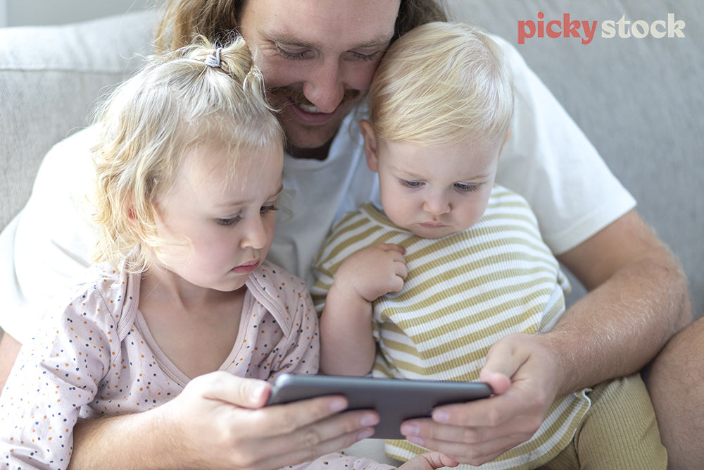 Landscape image of two small children sitting on the couch with their father, looking at phone screen. Dad with a big smile on face, while kids watch looking interested. 