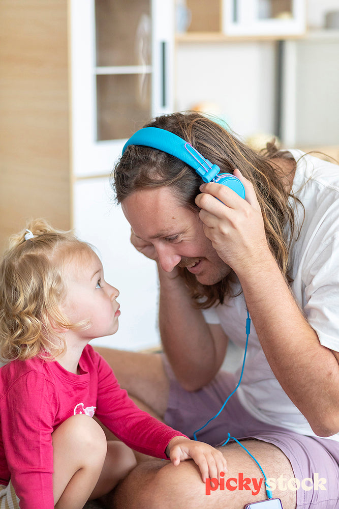 Small girl looking up at father who is wearing her blue headphones. Having a listen of her music. 