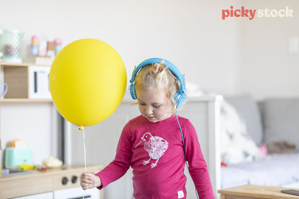 Landscape of small girl wearing a pink top with a kiwi illustration. Child is wearing bright blue headphones holding a bright yellow helium balloon. 
