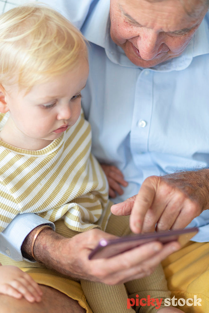 Portrait image of small boy sittng on the couch next to grandfather who is holding  a smartphone. Child is looking down at the screen. 