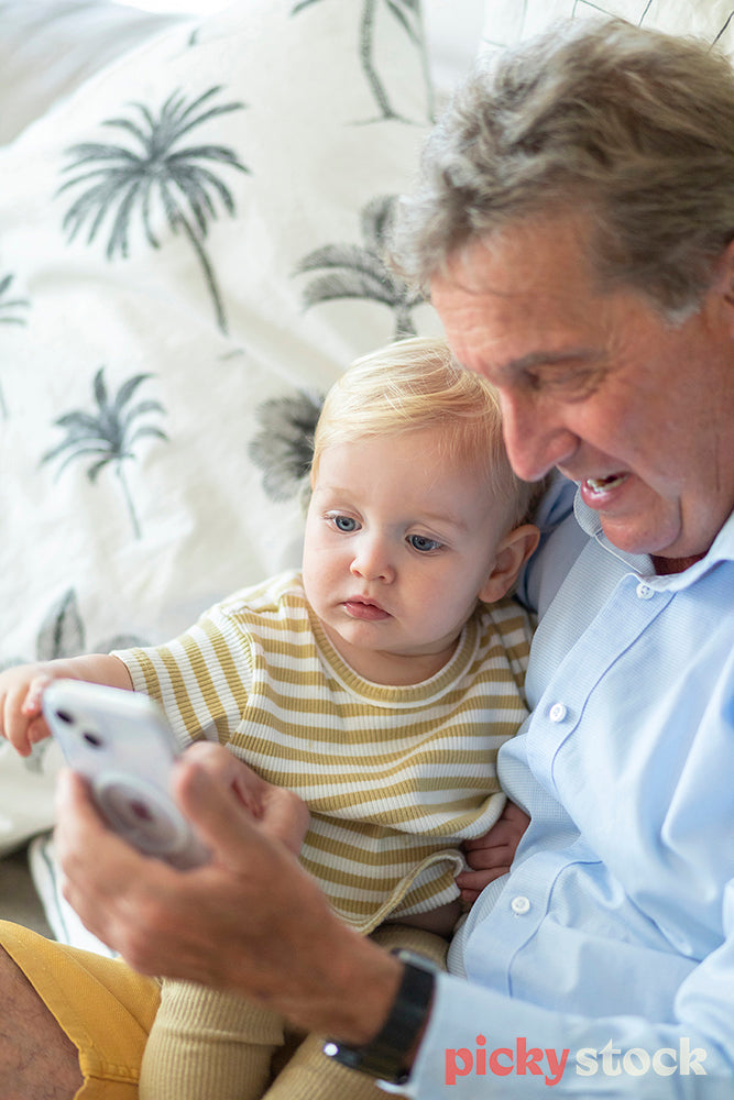 Portrait image of small boy sittng on the couch next to grandfather who is holding  a smartphone. Child is pointing at the screen. 