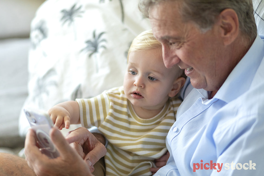 Landscape of small boy sittng on the couch next to grandfather who is holding  a smartphone. Child is pointing at the screen. 