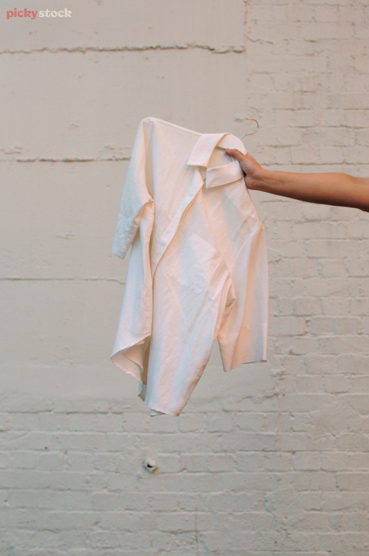 Portrait close-up of a tanned hand holding a small white shirt and clothes hanger at an angle against a whitewashed brick wall.