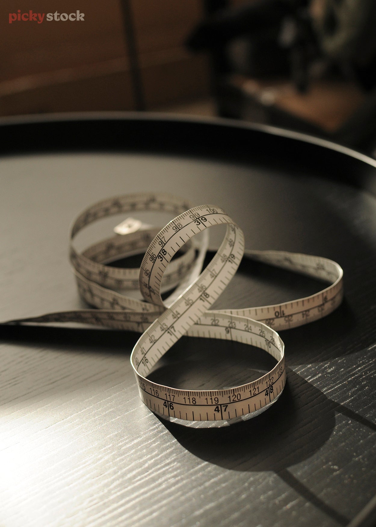 Portrait close-up of a fabric tape measure coiled atop a black wooden table the light casts black serpentine shadows around the surface of the table.