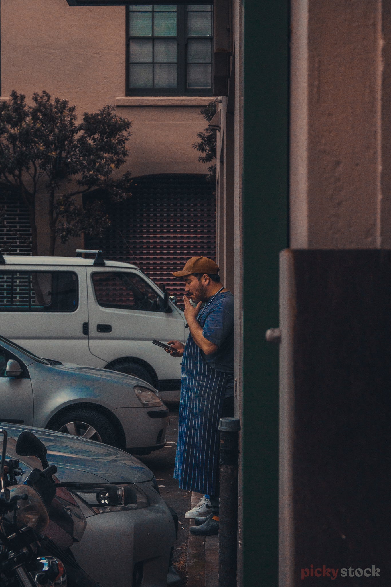 A chef takes a smoke break in a brown hat and striped apron while checking his phone. Pink concrete builnds and parked cars clutter the photo.