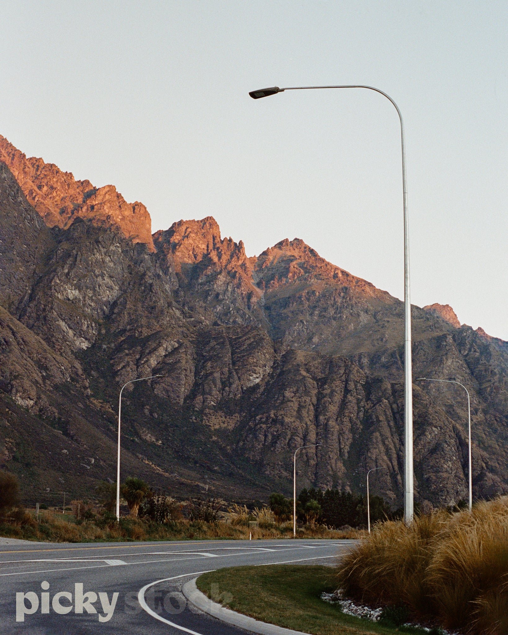 A portrait image of the Remarkables mountain range, with a road in the foreground. Large street lights hero the image, and are seen down the road. The road has a white line in the middle with flush strip. Mountain is lit with a soft golden light, early evening. Pastel sky. 