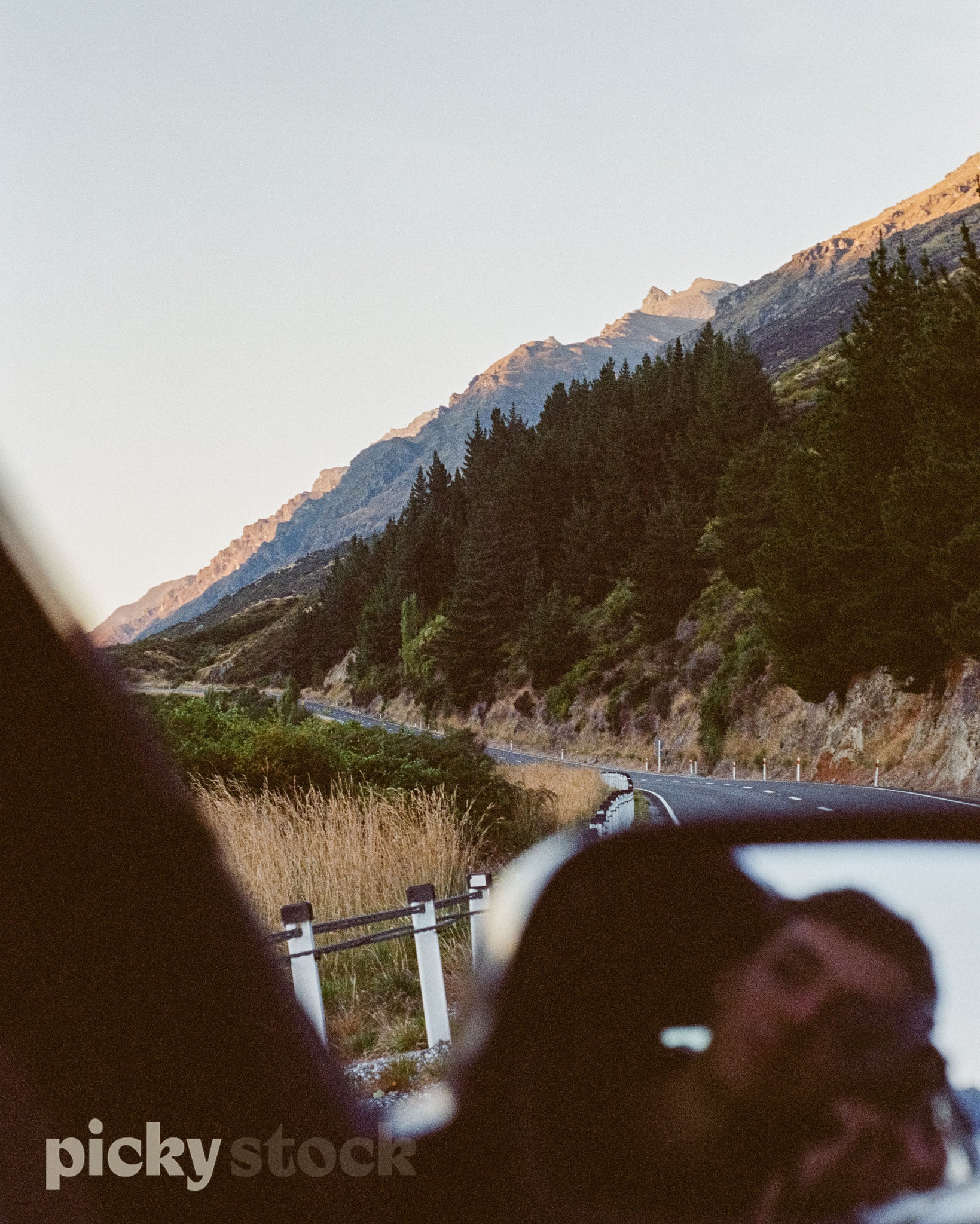 Man holding a camera, reflection of camera and face. Brown hair, grainy and soft focus. Car is driving on a rural road close to the mountain range. Green pine trees and mountain cliffs in the background. Road is well sealed with a barrier on each side. 