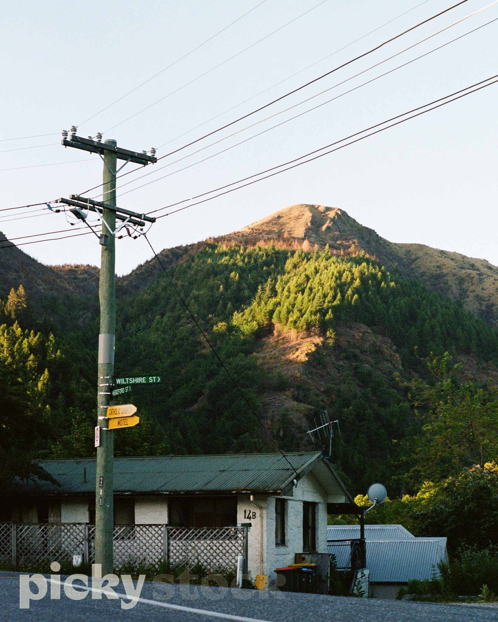 Portrait scene of a New Zealand Street, with a large power pylon and a bunch of yellow and green street signs pointing in various directions. Power lines running across the image. A small single unit yellow house with a green room, with the number 14B noted. Behind the house is a mountain range with dense pine trees scattered and grouped throughout. Golden light touching the mountain peak.