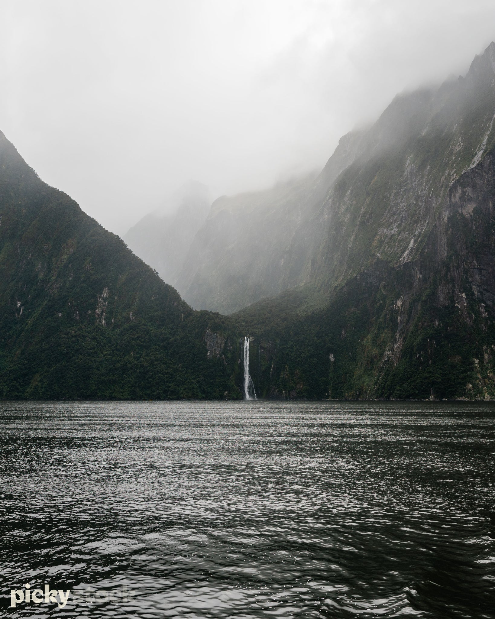 Portrait view of Milford Sounds, view from a boat. Foggy day, with dense fog low over the valley. Large cliffs in background. Water is dark and green. Image is moody. Waterfall is in the middle of the image.