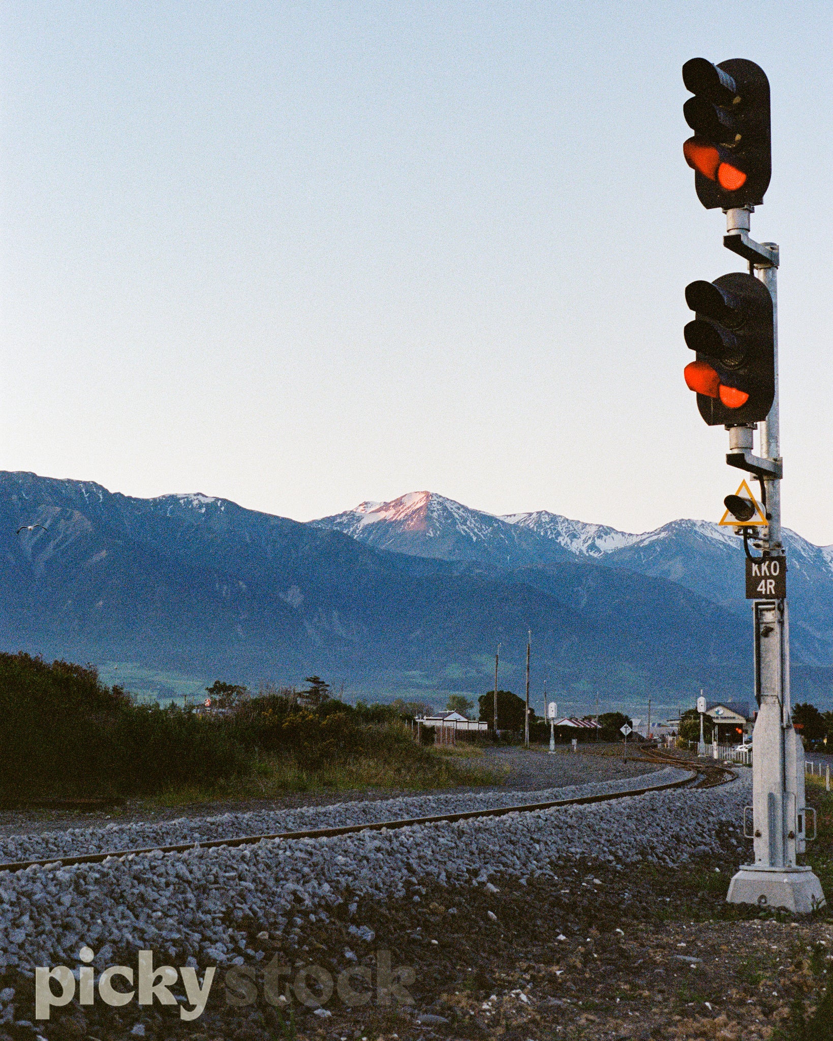Portrait image of a railway track. Two traffic light sets both showing a red signal. Background shows a view of a snow capped Mount Cook. View from summertime