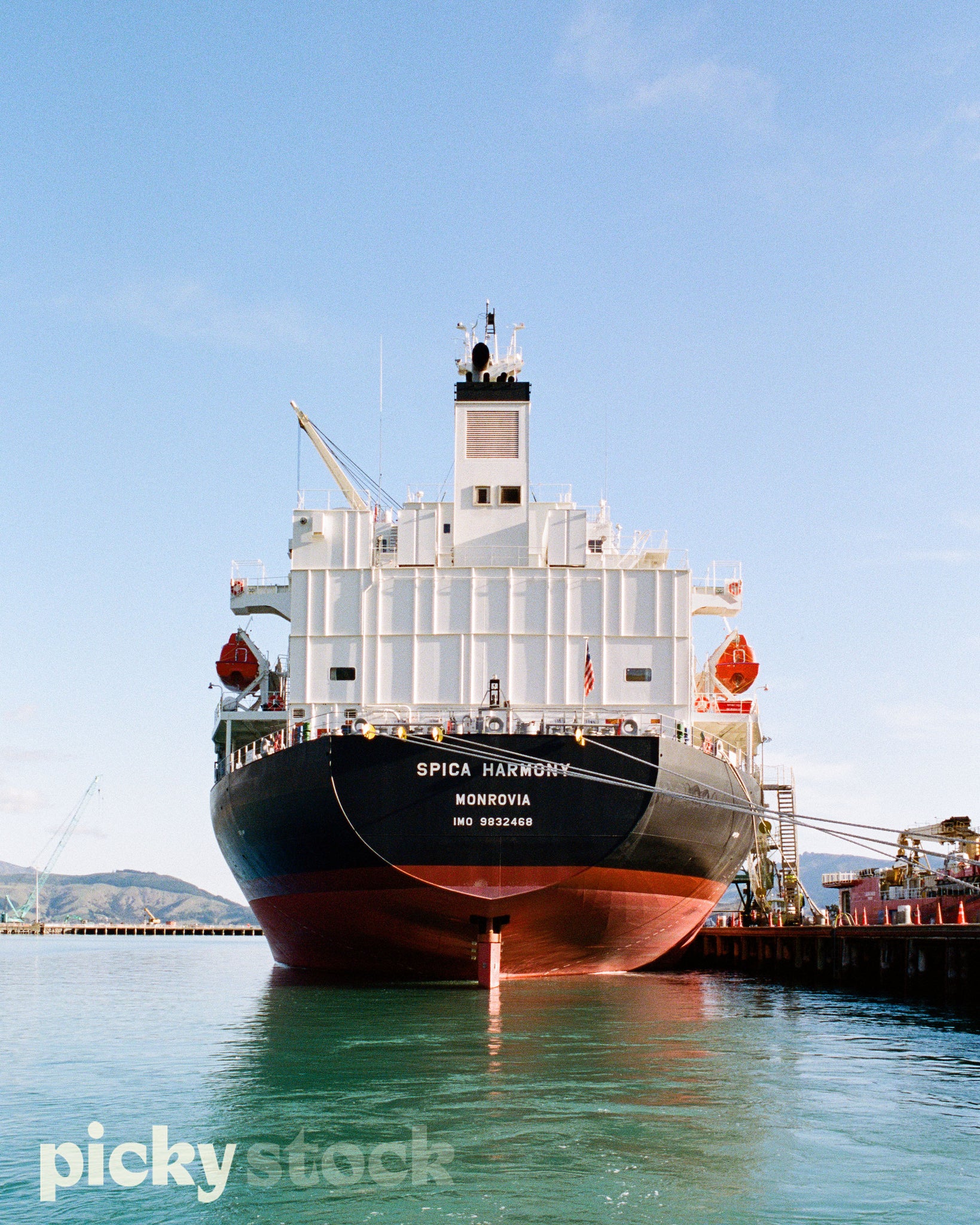 Portrait image of a large container ship docked at a port. Line holding boat to wharf. Wharf is to the right side of image. Container boat is has a red and black base with a white body and viewing platform. Sky is blue water is a turquoise green.