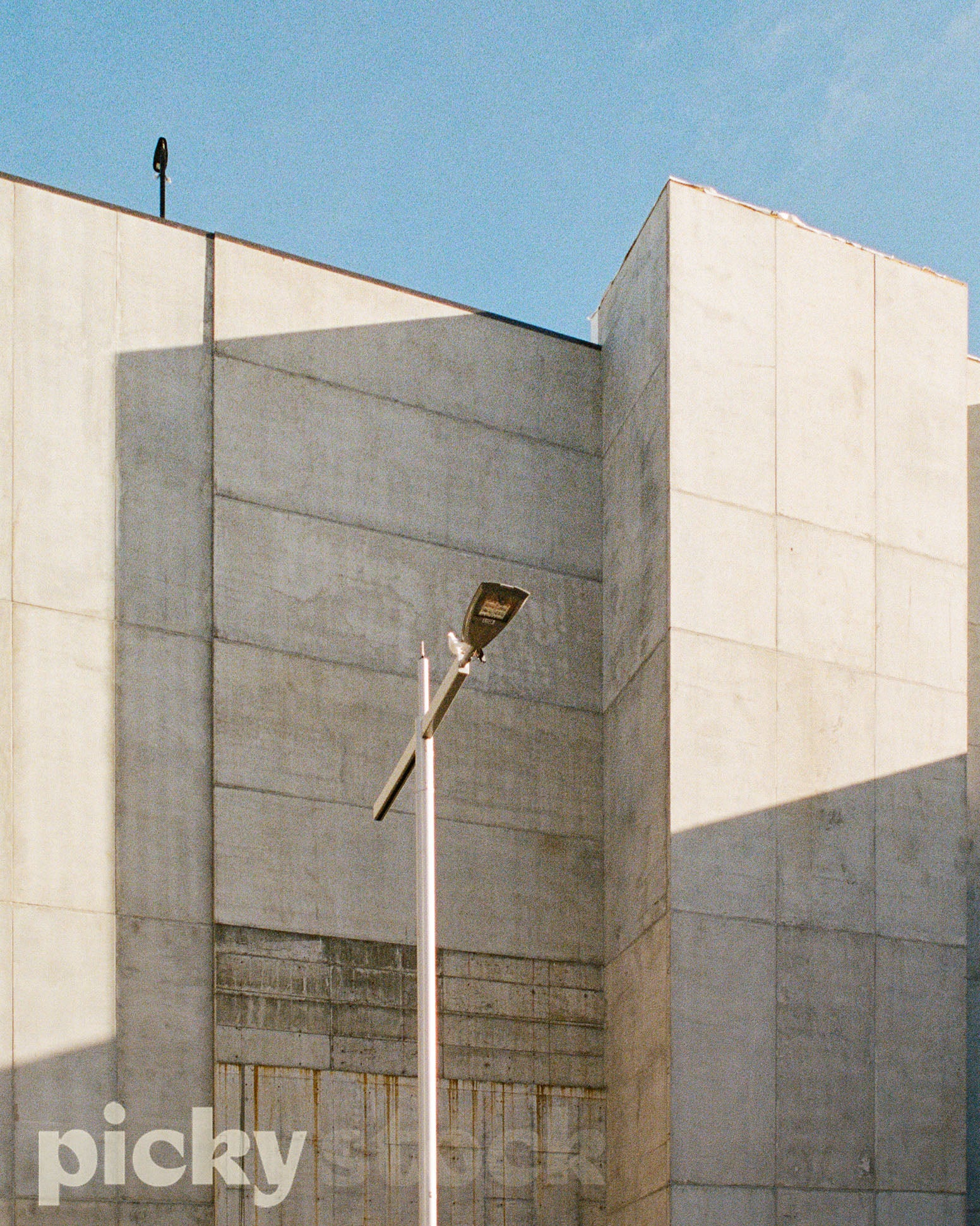 Giant concrete exterior of a car park building. Concrete is a rendered white in large chunks. Sky is blue. Large lamppost middle of frame.